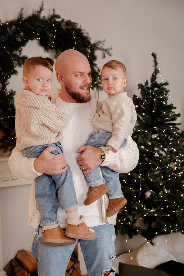Vater mit zwei kleinen Kindern auf den Armen neben einem beleuchteten Tannenbaum
